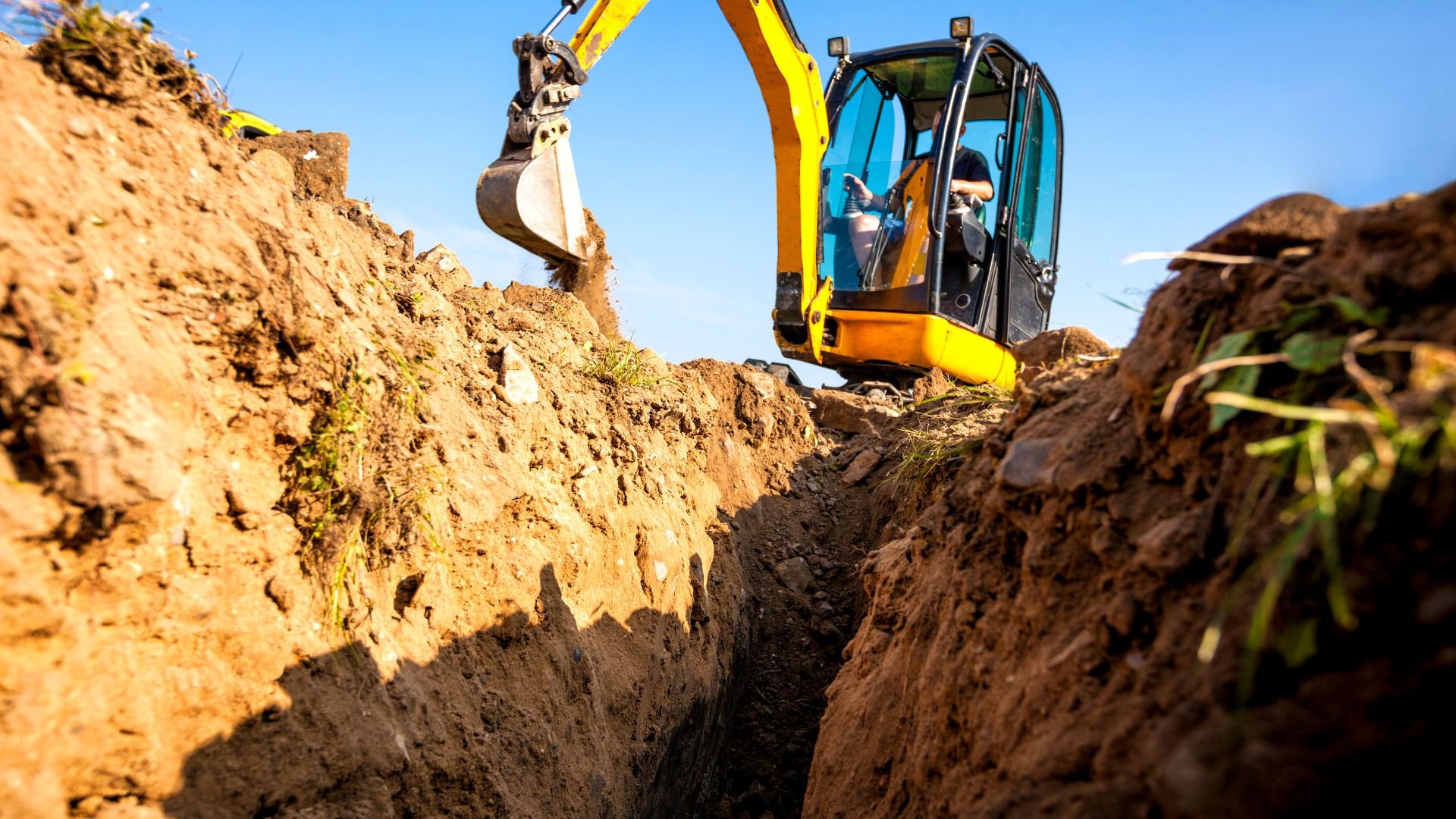 Yellow excavator digging deep trench in dirt under clear blue sky