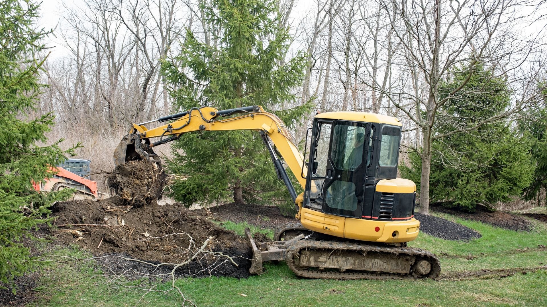 Yellow excavator digging in wooded area with trees and grass