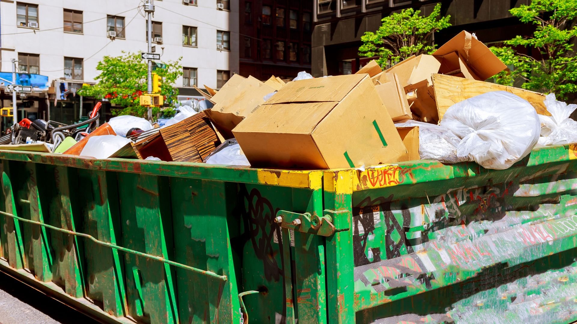 Green dumpster filled with cardboard boxes and white trash bags in urban setting